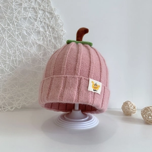 Close-up of pink knit baby hat with green leaf detail and small 'Baby' label on white stand
