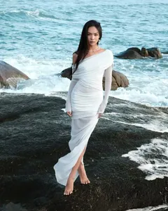 Close-up of white off-shoulder mesh maxi dress with ruching detail against ocean rocks