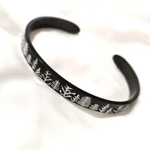 Detail of a black open cuff bracelet featuring a minimalist white leaf pattern on metal