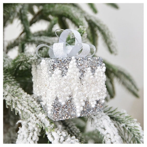 Silver sequin gift box ornament with white ribbon hanging on a snowy Christmas tree branch
