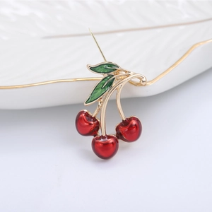 Close-up of red enamel cherry brooch with green leaves and gold stem on white ceramic