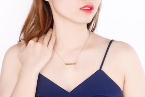 Close-up of a gold bar necklace featuring a small blue gem and heart engraving on a model