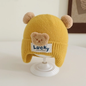Detail of yellow knit toddler hat with two brown pom-poms and a plush bear patch