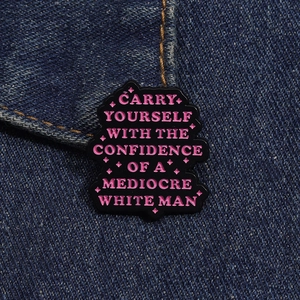 Detail of black enamel pin with pink text carry yourself with confidence on denim fabric