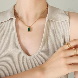 Golden necklace with square emerald green pendant shown on a model with gold rings