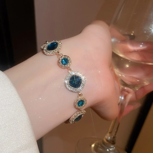 Nahaufnahme eines Goldarmbands mit tiefblauen Edelsteinen am Handgelenk mit einem Glas