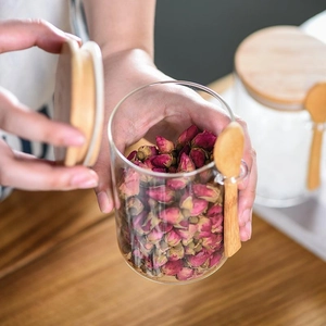 A round glass storage jar with a bamboo lid and spoon, held by hands in a kitchen setting