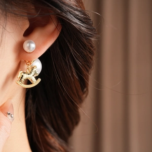 Detail view of pearl stud earring with golden rocking horse pendant on model