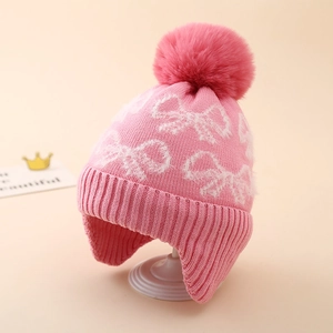 Pink knit trapper hat for girls with white bow patterns and a soft faux fur pompom