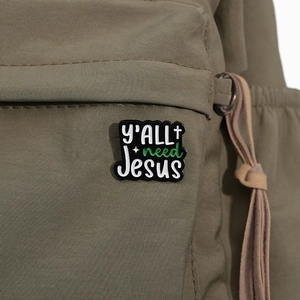 Close-up of 'Y'ALL NEED JESUS' enamel pin badge attached to a green canvas backpack
