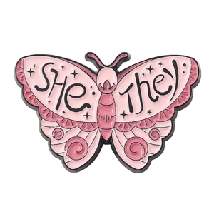 Detailed view of pink enamel butterfly pin featuring She and They pronoun identifier text