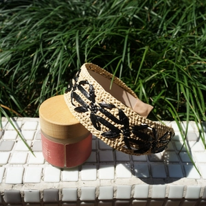 Detail of beige raffia headband with black floral embroidery on a white tile surface