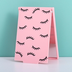Pink foldable makeup mirror with eyelash pattern, standing on a white surface