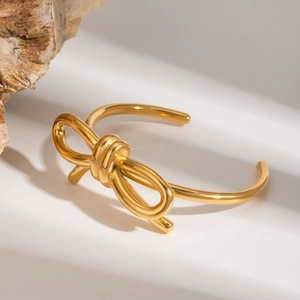 Detail of a gold-plated stainless steel bow bangle on a minimalist white stone surface