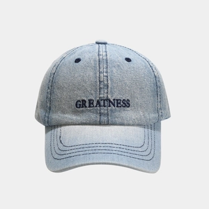 Detailed close-up of GREATNESS embroidery on light blue washed denim baseball cap