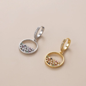 Goldene und silberne Huggie-Ohrringe mit bunten Zirkon-Anhängern auf beige Hintergrund