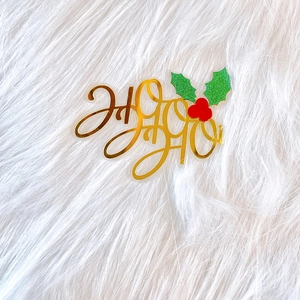 Close-up of golden Ho Ho Ho acrylic cake topper with green glitter holly leaves