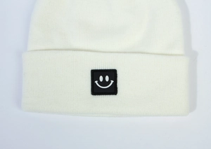 Close-up of white knit beanie featuring a square black smiley face patch detail