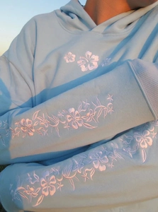 Detaillierte Nahaufnahme weißer Blumenstickerei auf einem hellblauen Kapuzenpullover im Licht