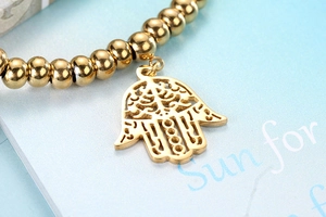 Detail of golden beaded bracelet with Hamsa hand charm on a soft blue display card