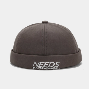 Detail of dark brown brimless docker hat featuring white 'NEEDS' embroidery on cotton fabric