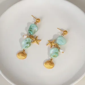 Golden earrings with green stones, pearls and starfish pendants on a beach background
