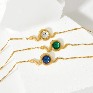 Three gold snake bracelets with green, blue, and clear gemstones on a white textured block
