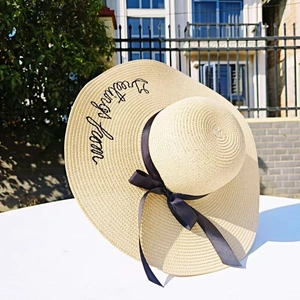 Detail of embroidered 'greetings from' text on a beige straw hat with a navy ribbon