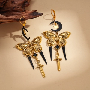 Golden butterfly earrings with black cone and sword charms on a minimalist wood background