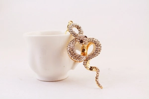 Detail of a sparkling golden snake brooch with red crystal eyes on a ceramic cup