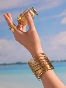Detail of layered gold wire bangles and sun ring set worn against a blue ocean view