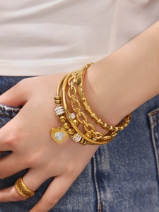 Detalle de pulseras doradas superpuestas con dijes de corazón y un anillo tejido en modelo