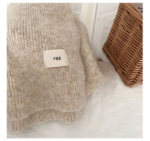 Close-up of soft beige wool scarf texture with 'egg' fabric label in a cozy home setting