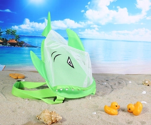 Green shark-shaped mesh beach bag for kids on sunny sand with blue ocean backdrop