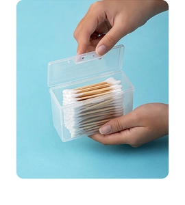 Hand holding a portable clear cotton swab storage box with wooden tips on blue background