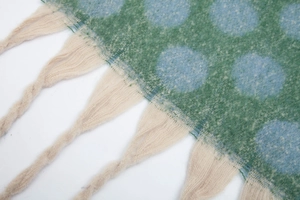 Dettaglio macro di una sciarpa in maglia con pois azzurri e nappe beige su fondo verde