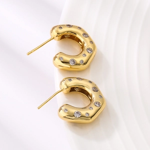 Gold C-shaped earrings with round clear stones on a white textured background