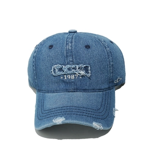 Detail of distressed denim baseball cap featuring white 1987 embroidery and ripped effects