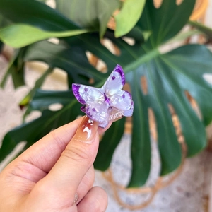 Chic purple butterfly hair clip with pearlescent finish held against tropical leaves