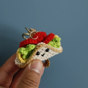 Handmade crochet taco keychain with a cute smiley face held in hand against blue background