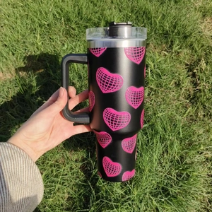 Close-up of 40oz black insulated car mug with pink disco heart patterns on grass