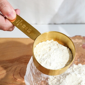 Hand holding a gold stainless steel 1/3 cup measuring spoon pouring flour into a bowl