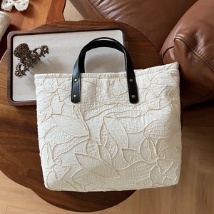 Quilted tote bag with leaf embroidery and black leather handles on a dark wood table