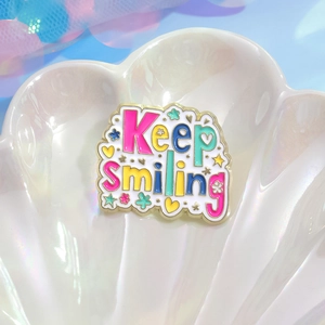 Keep Smiling enamel pin with colorful letters and gold metal base on a white shell plate