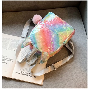 Rainbow sequin unicorn mini backpack with pink pompom charm on a book and grey sofa