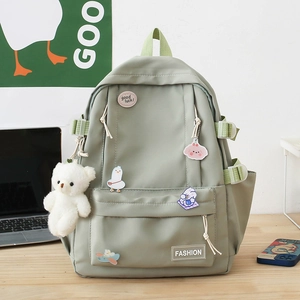 Pastel sage green school backpack with cute plush bear and cartoon pins in a desk setting