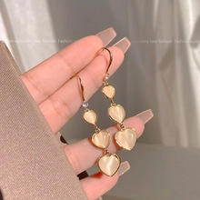 Detail view of long gold drop earrings with heart-shaped cat-eye stones held by hand