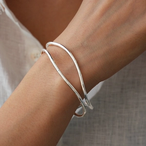 Close-up of a silver wavy double-layer open cuff bracelet worn on a wrist over grey linen fabric