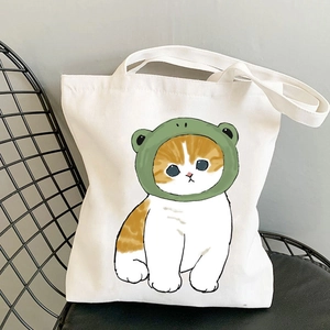 White canvas tote bag featuring cute orange cat with green frog hat print on metal chair