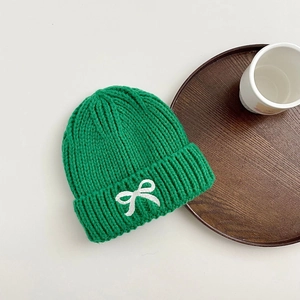 Close-up of green knitted beanie with white embroidered bow on a clean white table